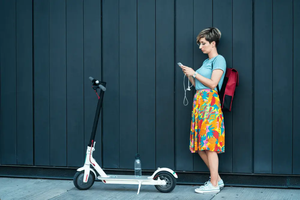A lady in sky t-shirt and multicolor skirt standing beside a white and black electric scooter
