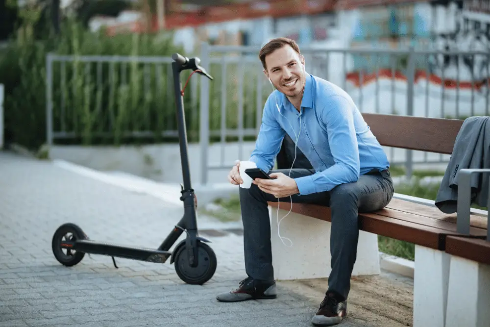 A guy with smiling face sitting beside an electric scooter holding a cup of coffee