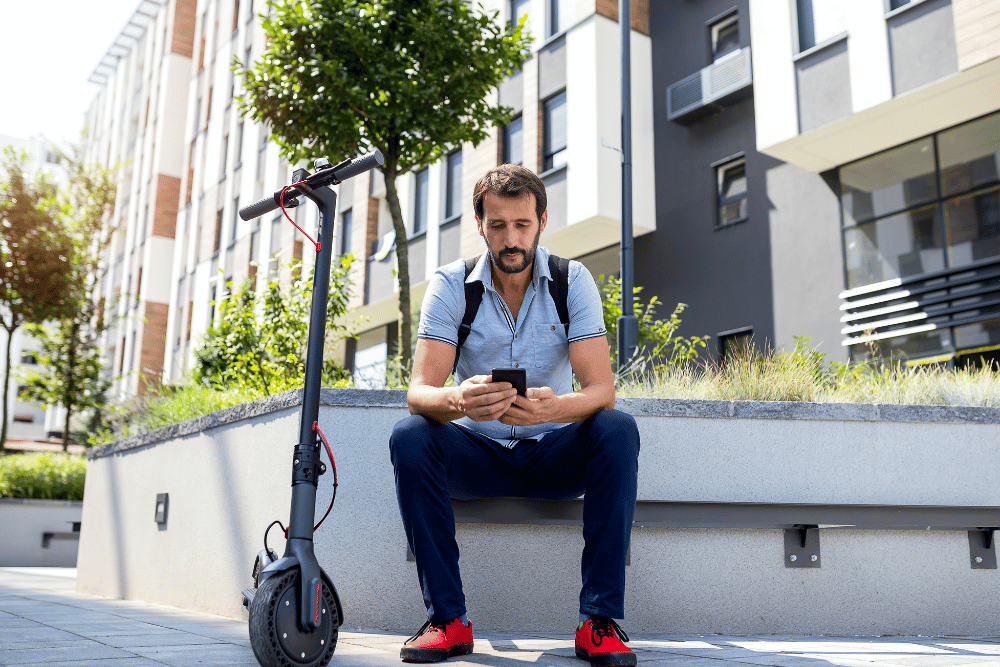 A man with back pack sitting on a road side along with an electric scooter
